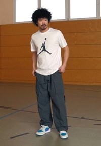 Hvid T-shirt med et sort Jordan-logo, grå cargo bukser og multicolore sneakers. Står på et gymnastikgulv med trævægge.