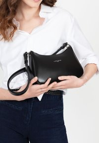 Sac à main en cuir noir, de forme courbée, avec une courte sangle réglable et un logo de la marque embossé à l'avant. Texture lisse.