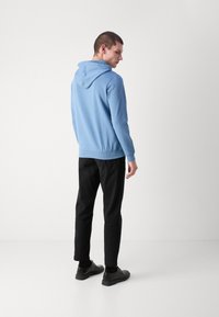 Camisola com capuz de tom azul claro, com cordão de ajuste, combinada com calças pretas e sapatos pretos. Tecido suave, design casual e silhueta ajustada.