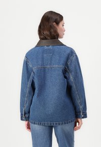 Denim jakna s temno rjavim ovratnikom, ohlapnim krojem in dolgimi rokavi. Ima vidne šive in gumbne manšete. Zadnji pogled.
