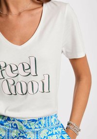 T-shirt bianca a maniche corte con scollo a V, con la scritta "Feel Good" in caratteri in grassetto e multicolori. Abbinata a pantaloni corti blu a fantasia.