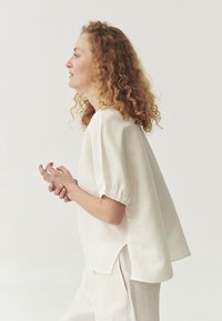 Cream, short-sleeve blouse with a loose fit and round neck. Features elasticized hems and side slits. Worn with matching light pants.