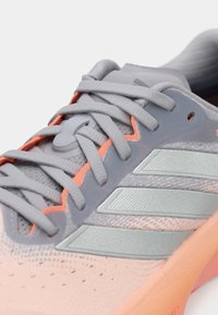 Sportschuh mit einem hellgrauen Mesh-Obermaterial, silbernen Akzenten und einer orangen Sohle. Verfügt über strukturierte Details und flache Schnürsenkel.