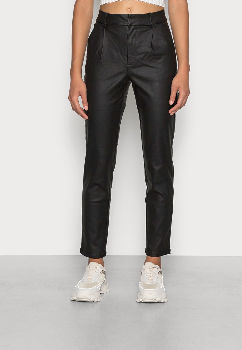 Pantaloni in pelle nera a vita alta per donna abbinati a scarpe da ginnastica bianche e spesse e a un top corto bianco, in piedi contro uno sfondo semplice.