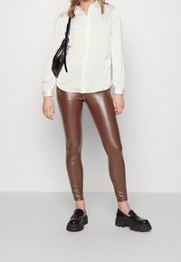 Brown faux leather leggings paired with a white button-up shirt and black platform loafers. Model holds a small black handbag.