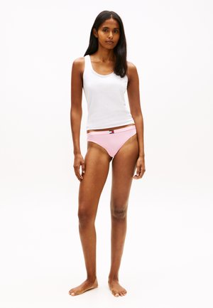 Kvinde står barfodet iført en hvid ærmeløs tanktop og lyserøde bikini-underbukser med et lille logo på linningen.