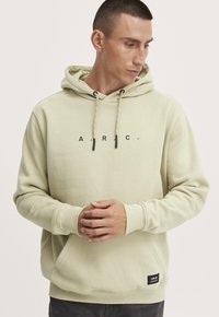 Lysegrøn hoodie lavet af blødt stof, med en frontlomme, snører med sorte ender og et minimalistisk logo trykt på brystet.