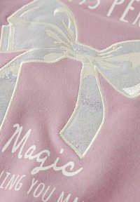 Tissu rose clair avec un nœud brodé blanc et des accents étincelants. Le texte dit "Magie" dans une police en cursive. Texture douce.