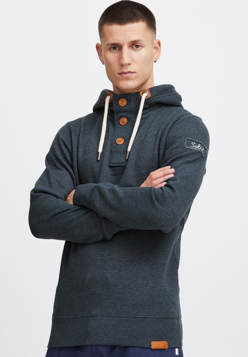 Sudadera con capucha en gris oscuro, con cuello abotonado, cordones en color crema, acentos en color beige y un parche de logo en la manga. Tejido suave y corte casual.