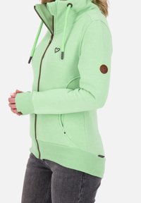 Sudadera con cremallera de color verde claro y alto cuello, con un logo de corazón, cremallera y detalles en marrón contrastante, y puños acanalados.
