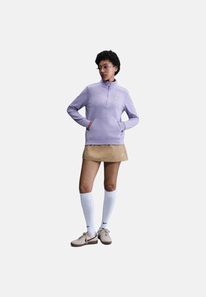 Sudadera de media cremallera lila con un bolsillo delantero, combinada con una falda beige y calcetas blancas hasta la rodilla; modelo lleva zapatos beige.