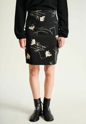 Mujer con falda negra hasta la rodilla con motivos florales beige, camiseta negra de manga larga y botas negras al tobillo, de pie frente a un fondo claro.