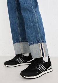 Sneakers neri con strisce bianche, abbinati a jeans in denim blu con orlo ripiegato e fodera interna a contrasto. Suola morbida, design minimalista.