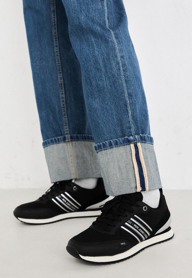 Baskets noires avec des rayures blanches, assorties à un jean en denim bleu avec un revers replié et une doublure intérieure contrastante. Semelle souple, design minimaliste.