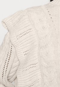Détail d'un pull en maille couleur crème avec des motifs torsadés et de dentelle mettant en valeur la texture et le design au niveau de l'épaule.
