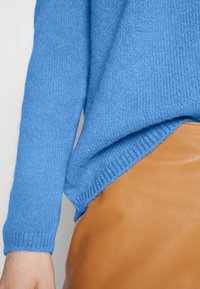 Maglione blu lavorato a maglia infilato in una gonna di pelle beige a vita alta con cuciture visibili in vita, su uno sfondo semplice.
