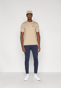 Camiseta de algodón beige con mangas cortas, con un pequeño logo; combinada con pantalones deportivos azul marino y zapatillas blancas; gorra beige en la cabeza.