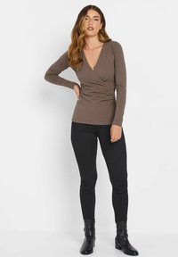 Brauner, langärmeliger Wickeloberteil mit tiefem V-Ausschnitt, tailliertem Design und weichem Material, kombiniert mit schwarzen Skinny-Jeans und Stiefeletten.