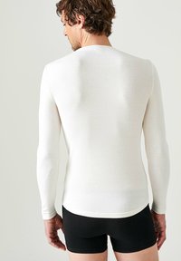 Camicia bianca a maniche lunghe con scollo rotondo, forma aderente e texture liscia, abbinata a pantaloni corti neri. Vista da dietro.