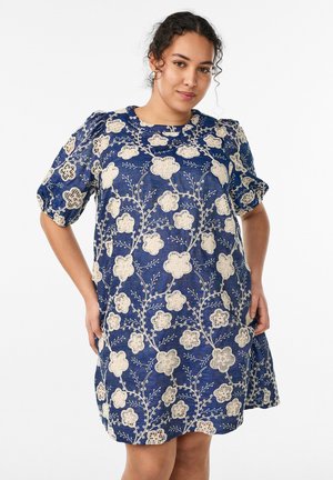 Žena v modrých šatách po kolená s nafúknutými rukávmi a béžovým kvetinovým krajkovým vzorom, stojaca so založenými rukami na bokoch pred bielym pozadím.