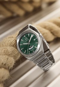 Montre-bracelet Timex en métal argenté avec cadran vert, affichage de la date et fonction GMT reposant sur une corde épaisse beige.