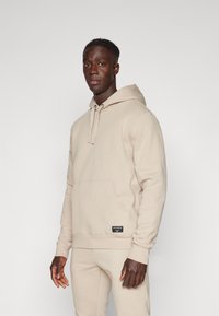 Sweatshirt capuz bege em mistura de algodão suave, com bolso frontal, punhos canelados e uma pequena etiqueta da marca na barra.