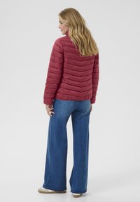 Giacca puffer a forma di rombo rossa con colletto alto, abbinata a jeans blu a gamba larga. Tessuto testurizzato, finitura liscia e dettagli hardware minimi.