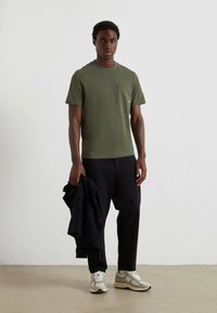 T-shirt à manches courtes vert olive avec une poche poitrine, associée à un pantalon sombre et des baskets grises, accompagnée d'une veste noire. Design simple.