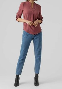 Blusa color ruggine con scollatura a V e maniche arrotolate, abbinata a jeans a vita alta di colore blu chiaro e stivali neri alla caviglia. Texture liscia, stile casual.