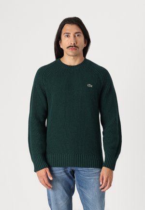 Groene geribbelde gebreide trui met een ronde hals en lange mouwen. Bevat een klein geborduurd logo op de linkerborst. Zachte textuur.