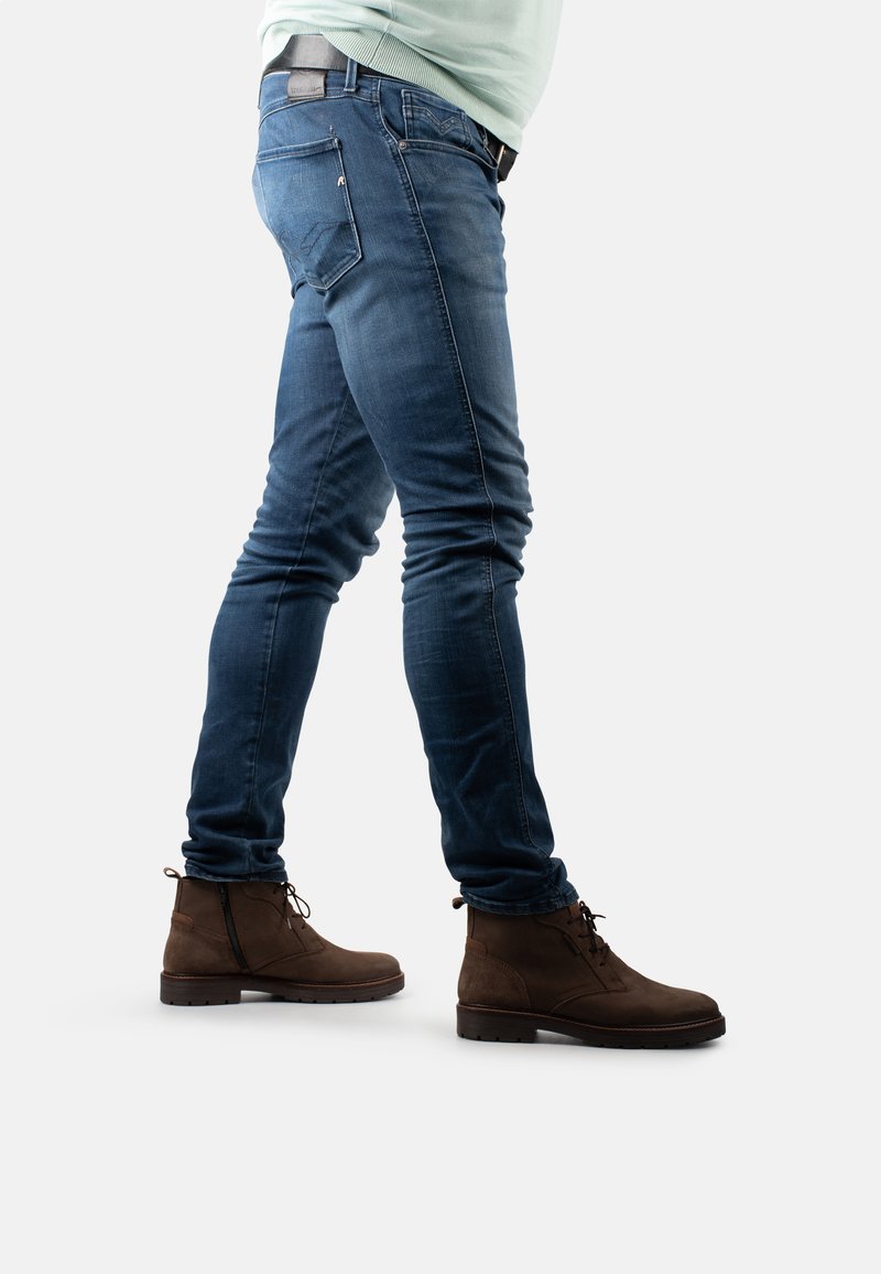 Dunkelblaue, enganliegende Jeans mit glatter Textur, ausgestattet mit einem Design für eine Gesäßtasche. Tragen mit braunen Wildleder-Stiefeletten und einem hellgrünen Oberteil.
