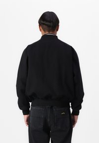Black bomber jacket with a rounded collar, elastic cuffs, and waistband. Worn over dark jeans with visible stitching and logo on the back pocket.