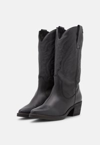 Par de botas vaqueras de cuero negro hasta la mitad de la pantorrilla con costuras decorativas, punteras redondeadas, lengüetas y tacones cuadrados sobre un fondo blanco.