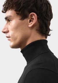 Černý rolák s ribbovaným límcem a hladkým povrchem, předvedený na profilu modelu proti jednoduchému bílému pozadí.