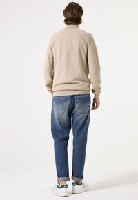 Beiger Pullover mit hohem Kragen und Rippendetails, kombiniert mit hochgekrempelten blauen Jeans und weißen Sneakers mit grünen Akzenten.