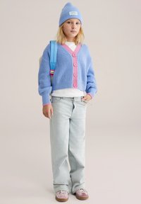 Cardigan azzurro chiaro lavorato a maglia con bottoni rosa, camicia bianca sotto, jeans larghi azzurro chiaro, sneakers viola e rosa, e berretto blu.