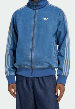 Man draagt een blauw denim Adidas trainingsjack met witte drie strepen op de mouwen en een rits aan de voorkant, gecombineerd met donkerblauwe broek.