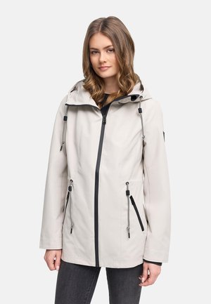 Femeie purtând o jachetă bej deschis cu glugă, fermoar negru și șnururi reglabile, stând într-un decor simplu.