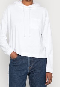 Hoodie bianco, corto, realizzato in morbido tessuto con tasca frontale e coulisse regolabile sul cappuccio, abbinato a jeans di denim blu.