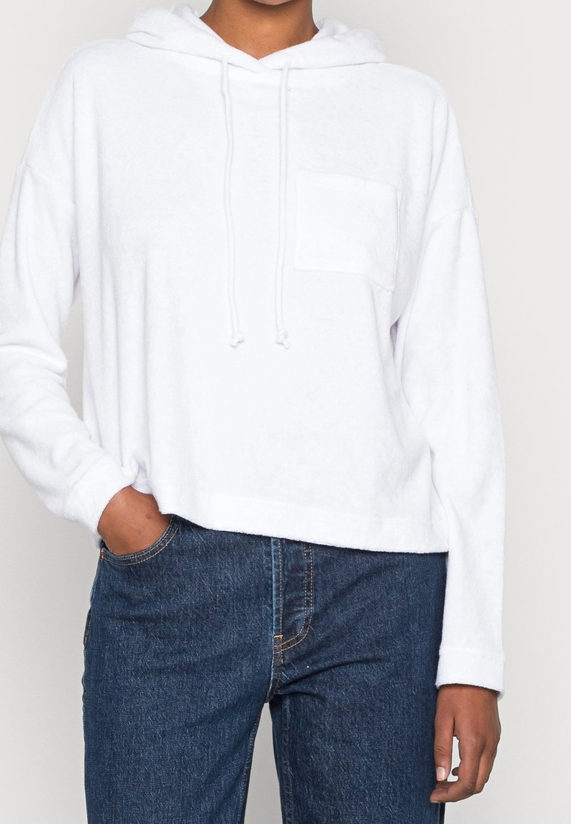 Hoodie bianco, corto, realizzato in morbido tessuto con tasca frontale e coulisse regolabile sul cappuccio, abbinato a jeans di denim blu.