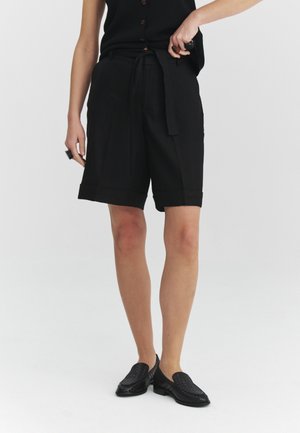 Mujer con pantalones cortos negros hasta la rodilla con cinturón atado y zapatos tejidos negros sin cordones, de pie contra un fondo blanco liso.