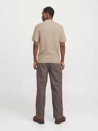 Mand iført en beige kortærmet striktrøje, grå bukser og brune sko, stående mod en ensfarvet hvid baggrund med ryggen vendt mod beskueren.