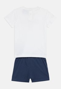 Λευκό μπλουζάκι μωρού με κοντά μανίκια και κουμπιά στο πίσω μέρος, συνδυασμένο με σκούρο μπλε σορτς με ελαστική μέση, τοποθετημένα επίπεδα σε λευκό φόντο.