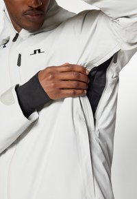 Casaco cinza claro, impermeável, com fecho frontal, punhos ajustáveis e bolsos laterais. Apresenta um forro de malha e um logótipo no peito.