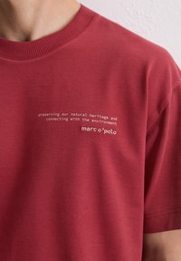 Rotes Baumwoll-T-Shirt mit rundem Ausschnitt. Auf der Vorderseite steht "Unser naturliches Erbe bewahren und mit der Umwelt verbunden sein."