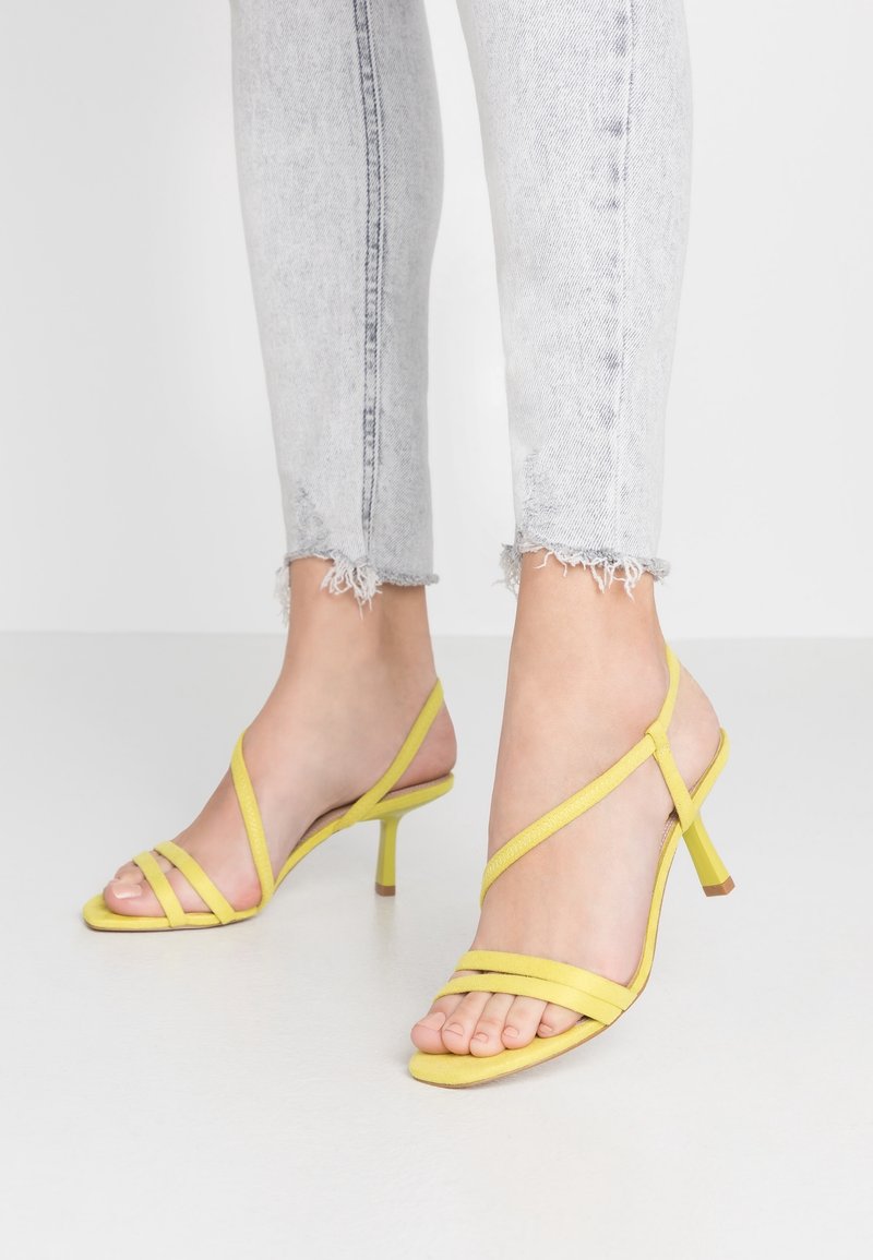 Sandales à talons hauts jaunes avec des lanières, un talon fin et un design ouvert sur l'avant ; fabriquées en matériau doux avec une texture lisse et peu d'accents.
