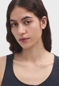 Zblízka na osobu s hnedými vlasmi strednej dĺžky, oblečenou v tmavomodrom topu bez rukávov. Pokožka má hladkú textúru a make-up je neutrálne ladený.