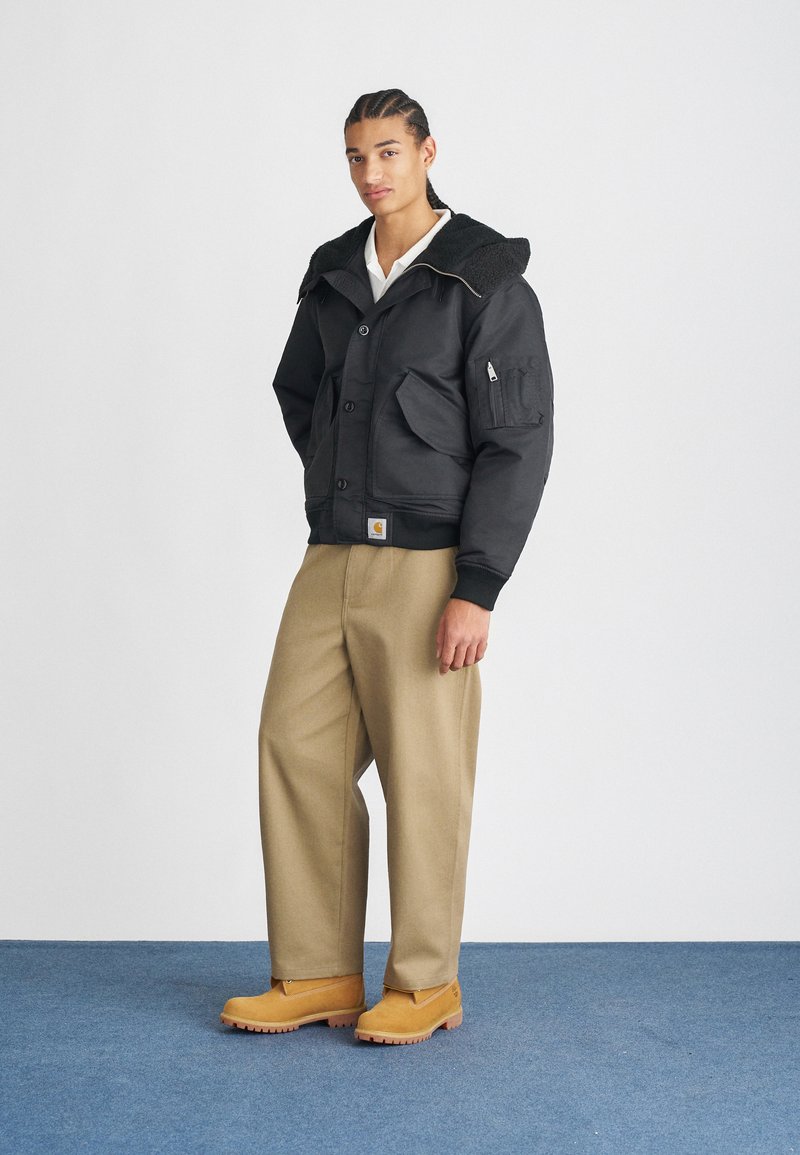 Veste bomber noire avec un col texturé, poches avant et fermeture à boutons. Associée à un pantalon kaki et des bottes beiges sur un sol bleu.