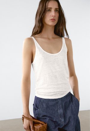 White, sleeveless tank top with a rounded neckline, made of lightweight fabric. Paired with navy blue, loose-fit trousers and a brown woven bag.