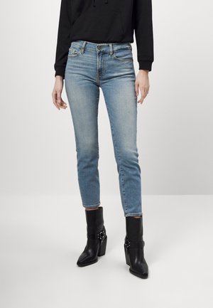 Femme portant un jean slim bleu clair, des bottines noires à talons hauts avec boucles et un haut noir à manches longues.